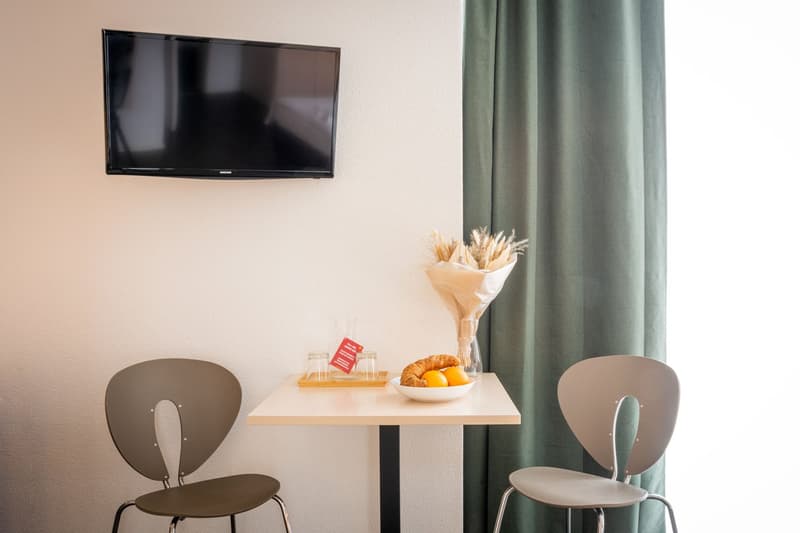 The image shows a hotel room with a flat-screen TV mounted on the wall, a small table with a bouquet of dried flowers, a plate of croissants and fruit, and two gray chairs. There appears to be a curtain on the right side of the room.