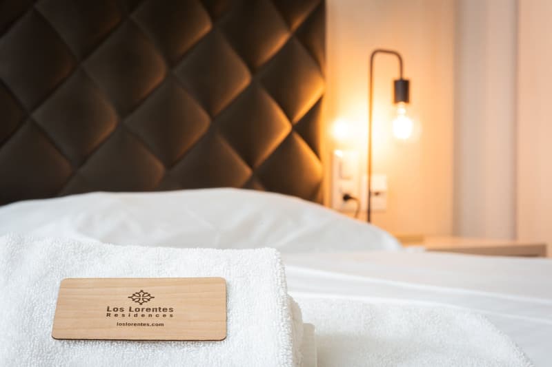 Cozy bedroom with textured wall panels, a white bedsheet, and a wooden business card for Los Lorentes Residences. A lamp provides warm lighting in the room.
