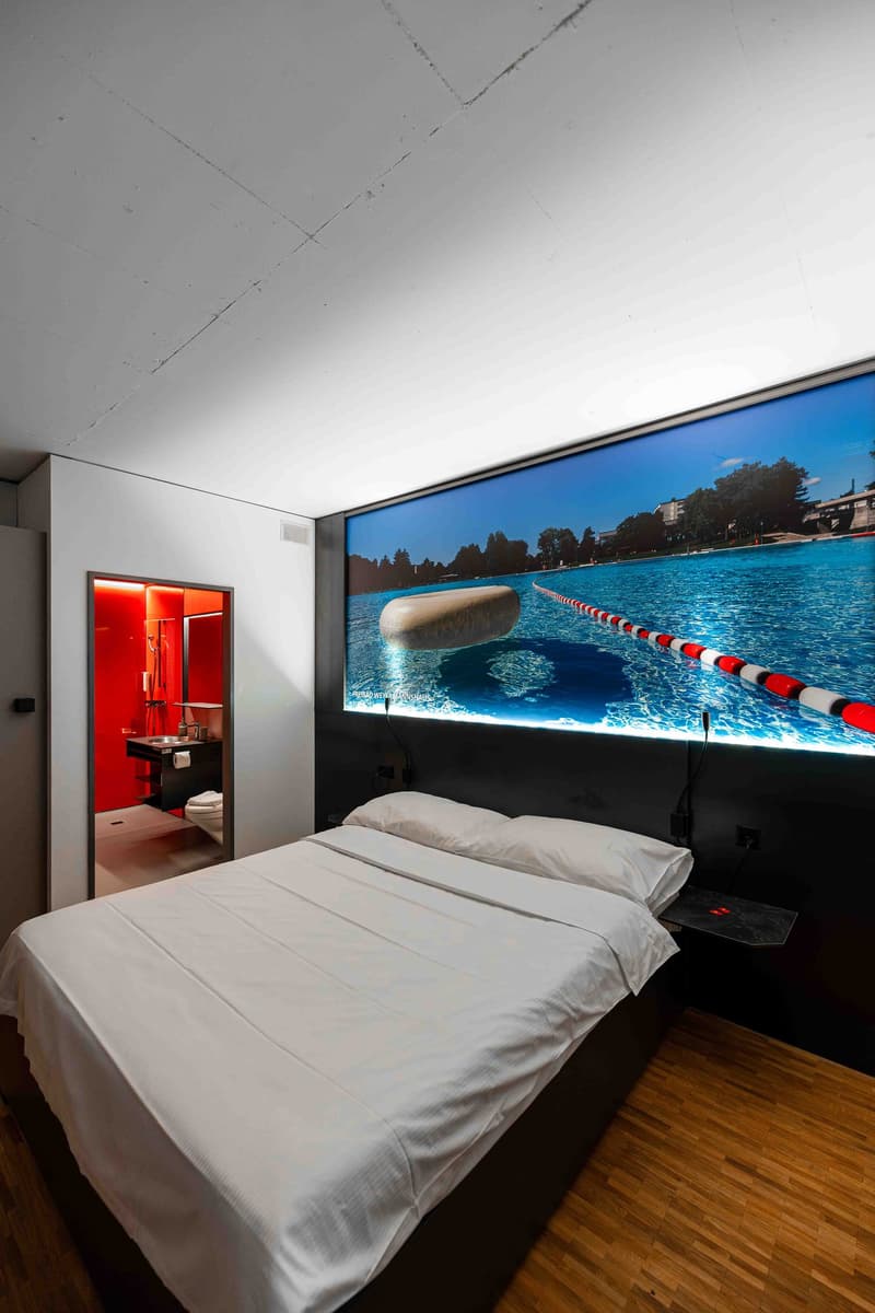 Bedroom with a bed, a large wall-mounted screen displaying a swimming pool, a wooden floor, and a red-themed bathroom visible through an open door.