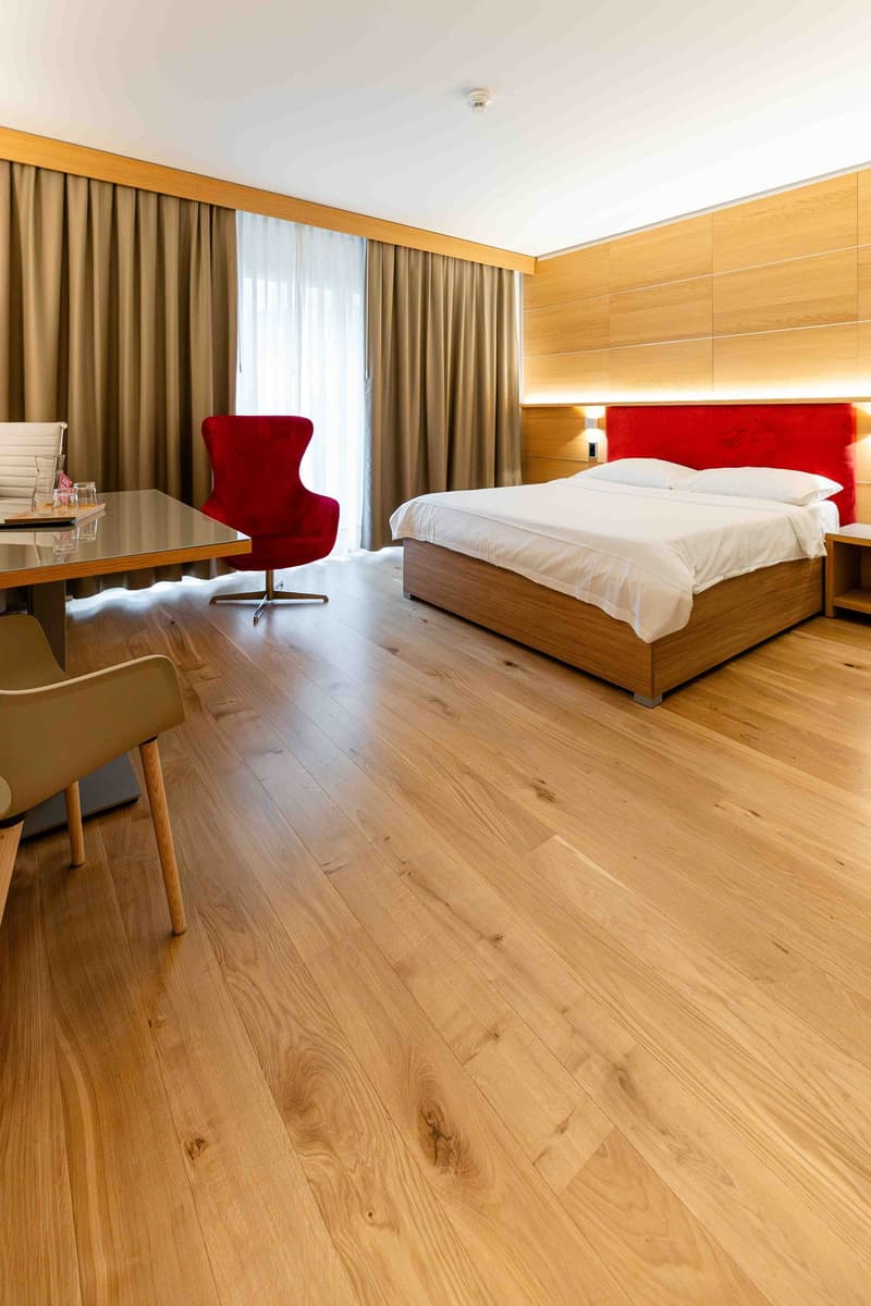 Bedroom with wooden floor, bed with white sheets, red headboard, red chair, desk with glass top, side table, curtains, and lamps