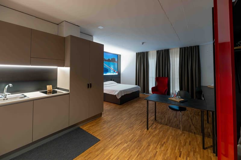 small room with a bed, desk, and a kitchen area with a sink and stove