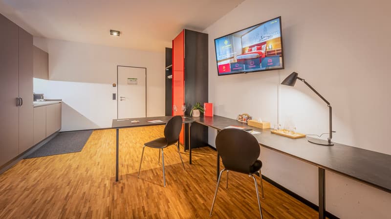 modern room with wooden floor, two desks, two chairs, TV, lamp, cabinet, door, kitchen area