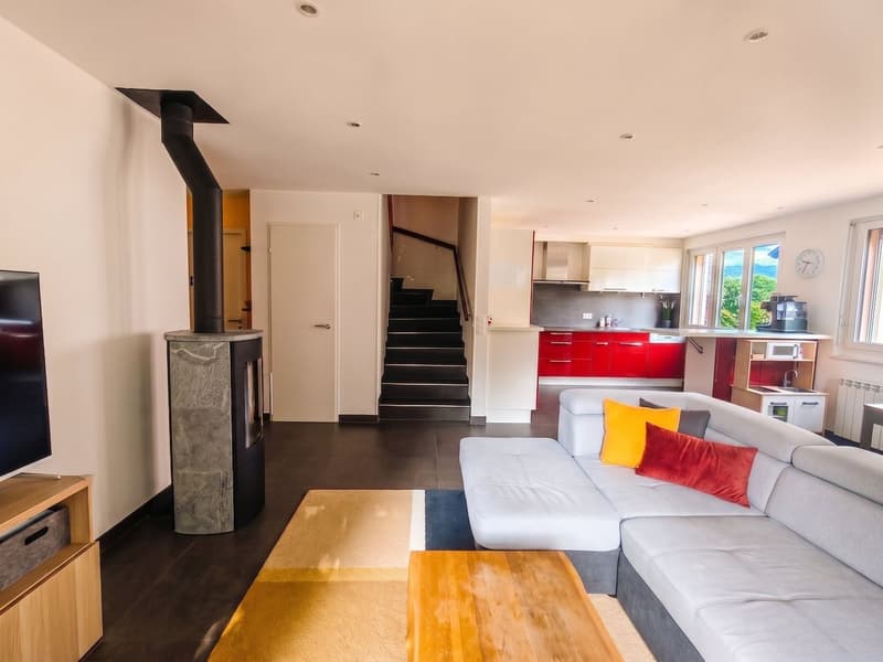 Salon avec vue sur l'escalier menant à l'étage, le poêle et la cuisine située en arrière-plan.
