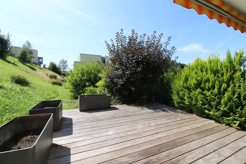 large wooden deck, green shrubs, 2 rectangular metal planters, beige awning