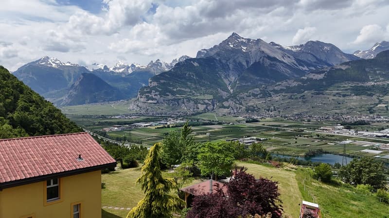 The image shows a stunning mountain landscape with snow-capped peaks in the background. In the foreground, there is a residential area with red-tiled roofs and lush greenery, including trees and a grassy area. The scene is framed by the surrounding mounta