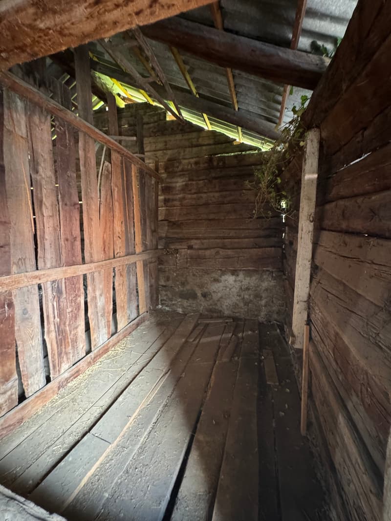 Dilapidated wooden interior with old, weathered boards and beams. The space appears to be a narrow, dimly lit hallway or passage with a dirt floor.