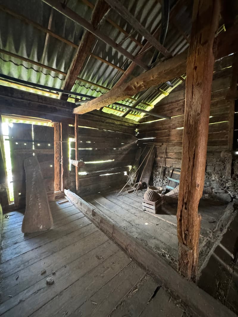 The image shows the interior of an old, rustic wooden barn or shed. The space is dimly lit, with wooden beams and a wooden floor visible. There are various tools and objects scattered around the space, suggesting it may have been used for storage or as a