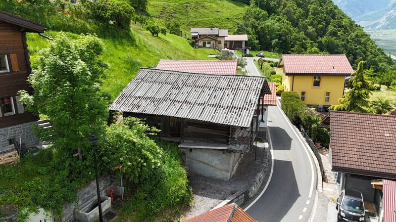 The image shows a scenic rural area with several houses and buildings nestled among lush green hills and trees. The buildings have traditional alpine-style architecture with sloped roofs and wooden construction. Some of the houses appear to be raised off