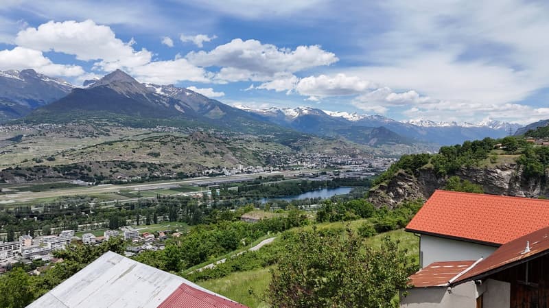 The image shows a panoramic view of a mountainous landscape. In the foreground, there are several buildings with red roofs. The landscape is dominated by lush green vegetation, including trees and fields. In the background, there are snow-capped mountains