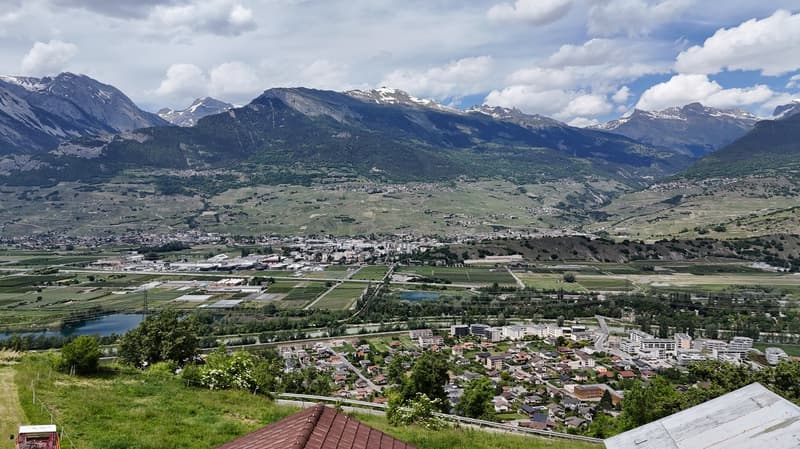 The image shows a panoramic view of a mountainous landscape. In the foreground, there is a small town or village nestled in the valley, surrounded by lush green fields and forests. In the background, towering snow-capped peaks rise up against a cloudy sky