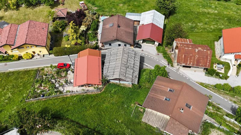 The image shows an aerial view of a residential neighborhood with various houses and buildings. The houses have red tile roofs and there is a garage visible in one of the buildings. The surrounding area is covered in lush greenery, including trees and gra