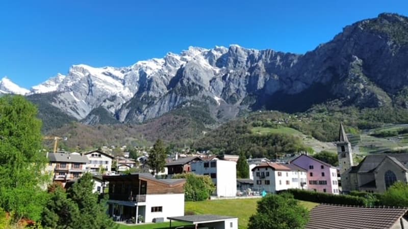 The image shows a picturesque mountain village nestled in a valley, with snow-capped peaks in the background. The buildings have a traditional alpine architecture style, with colorful facades and roofs. The landscape is lush and green, with trees and vege