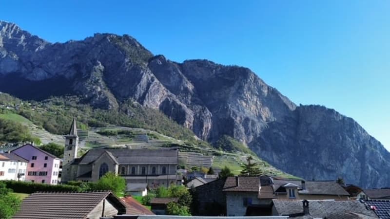 The image shows a picturesque mountain village nestled in a valley surrounded by towering rocky mountains. The buildings have traditional alpine architecture with sloped roofs, and some appear to be raised off the ground. The village is situated on a hill
