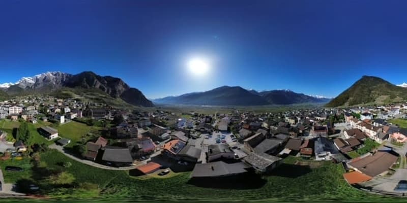This image shows a panoramic view of a mountainous town or city. The town is surrounded by mountains and has a mix of residential and commercial buildings with red roofs. The sky is clear and blue, with the sun shining brightly overhead.
