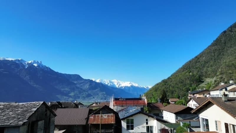 The image shows a scenic mountain landscape with a small town or village nestled in the valley below. The buildings have traditional alpine architecture with sloped roofs. The mountains in the background are snow-capped, indicating a high-altitude locatio