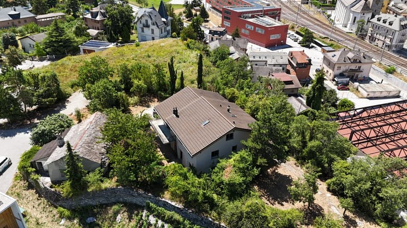Aerial view of a hillside neighborhood with various houses, including a 2-story brown house with solar panels on the roof, another with a red roof, and others of varying sizes and styles. There are multiple trees and greenery, along with an elevated red s
