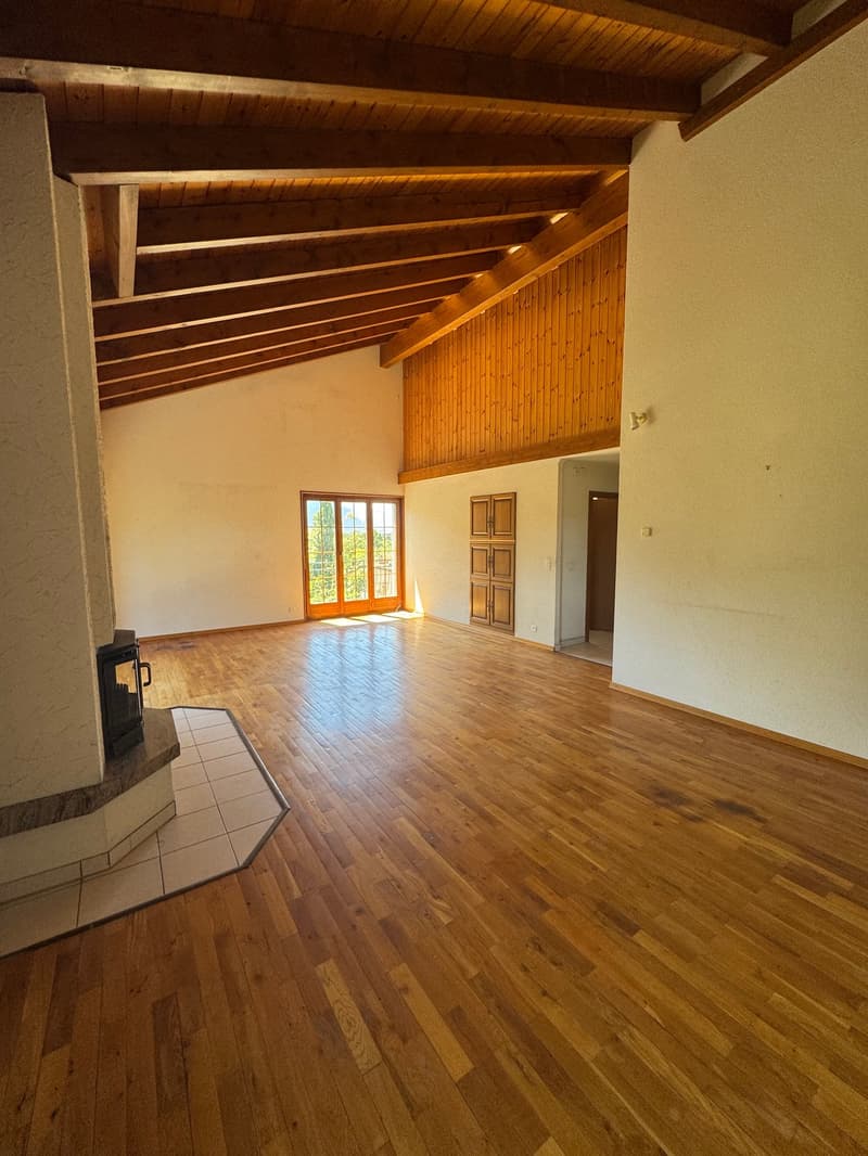 Empty room with hardwood floor, wooden beams, white walls, fireplace, glass doors with balcony, and a small balcony area with tiled floor. There are two wooden doors and a switch on the wall.