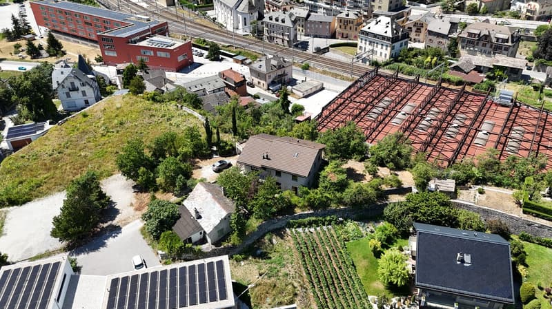 Various residential and commercial buildings in an urban area, multiple houses, red roof structures, a large red building with a metal framework, several smaller buildings, solar panels on buildings, greenery, and a driveway.