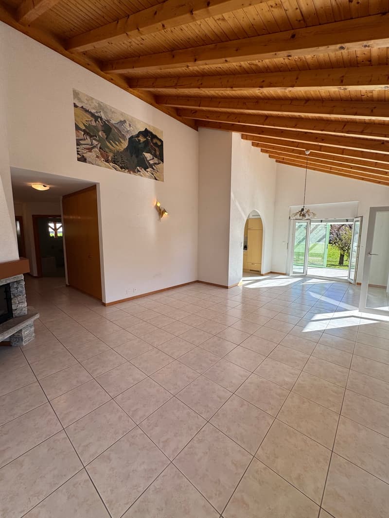 Empty living room with large glass doors, a fireplace, white tiled floor, wooden beams, paintings, recessed lighting.
