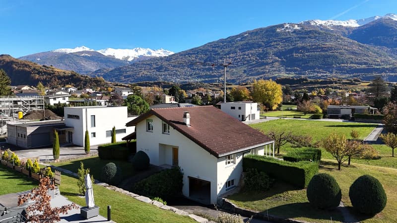 white house, brown roof, garage, well-manicured lawn, green landscape, mountains in the background