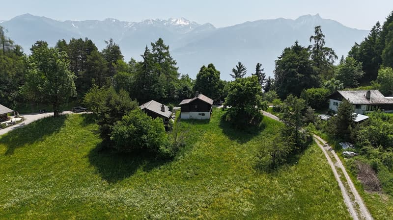 The image shows a scenic rural landscape with a mountainous backdrop. There are several small houses or cabins nestled among the lush green trees and vegetation. A dirt path or road winds through the grassy area in the foreground. The overall scene depict