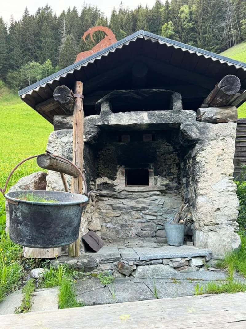 A rustic, stone-built outdoor structure with a thatched roof, likely used for cooking or heating. It has a large fireplace opening and several metal pots or buckets placed in front of it. The structure is situated in a grassy, forested area.