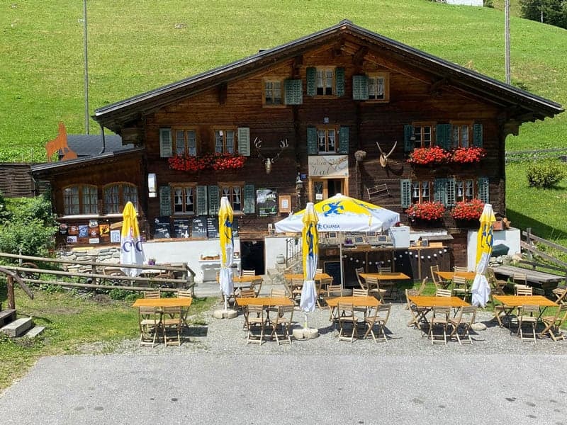 Traditional Swiss chalet-style building with wooden exterior, decorated with flower boxes and antlers. Outdoor seating area with yellow umbrellas and tables. Appears to be a restaurant or cafe in a rural, mountainous setting.