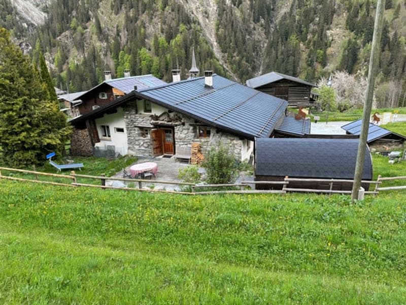 Two-story stone and wood house with a slanted metal roof, surrounded by a grassy field and trees. The house has a balcony or terrace on the upper level.