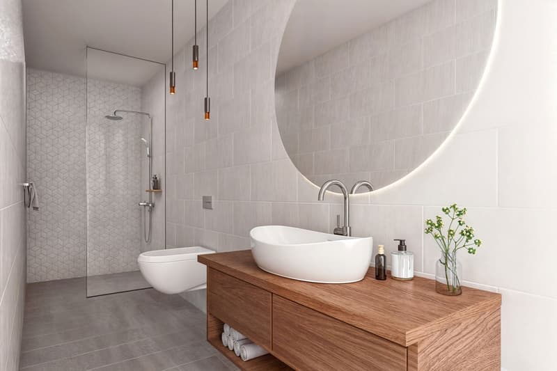 modern bathroom with a glass shower enclosure, toilet, large round mirror, wooden vanity, white porcelain sink, shower head, towels, lotion bottles, flowers