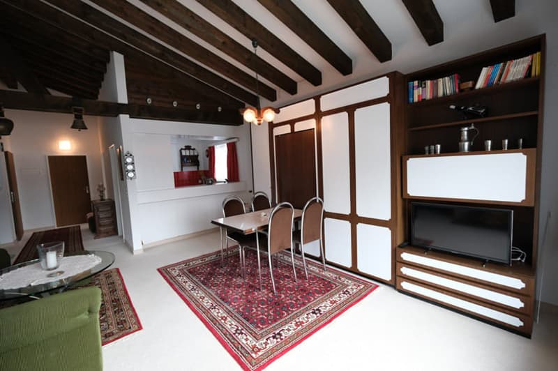 Dining room with wooden beams, dining table, chairs, rug, TV, shelves, and a green couch