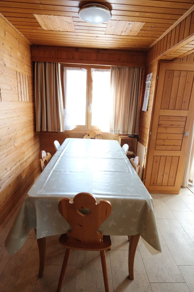Dining room with wooden walls, a long table, and chairs. A window with curtains and a ceiling light.