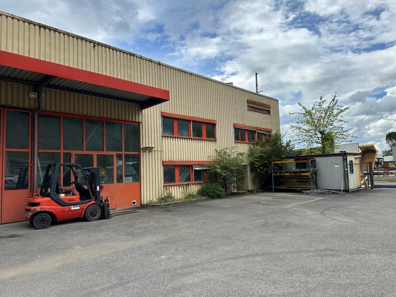 A large industrial building with a red and gray metal exterior, featuring large windows and a loading dock area. There is a forklift visible in the foreground, indicating this is likely a warehouse or manufacturing facility.
