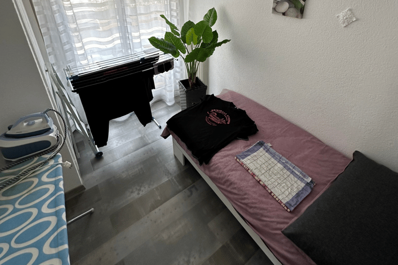 Small bedroom with bed, window, large plant, iron and ironing board, t-shirt on bed