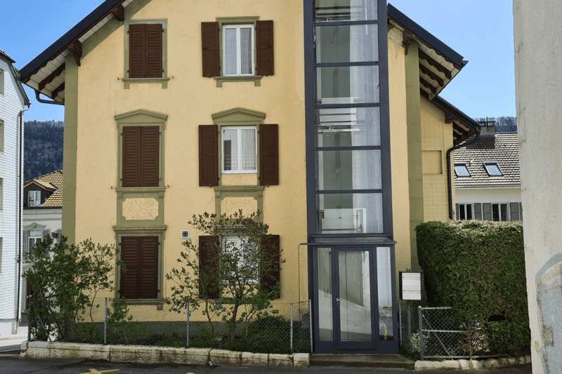 Yellow building with brown shutters, one glass elevator, entrance at the bottom, fence