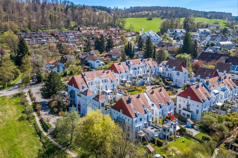 Aerial view of a residential area with multiple houses, some with red roofs, surrounded by trees and greenery. Some houses have balconies and raised ground floors.