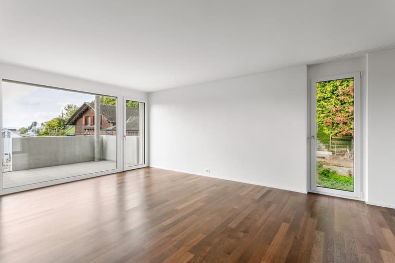 Spacious empty room with hardwood floors, large windows, double doors, sliding glass doors, balcony with a view of trees.
