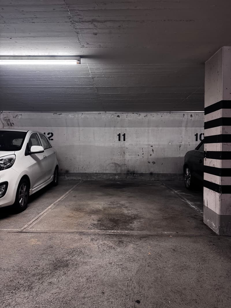 Underground parking area, two cars parked, concrete walls, numbers 10, 11, and 12 on the wall, fluorescent light on ceiling