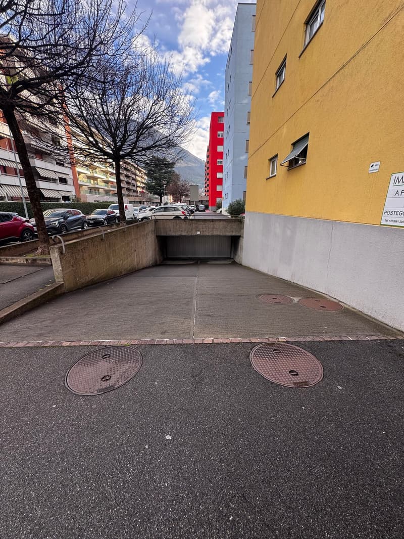 concrete paved parking lot, metal covers, garage entrance at the end, multiple buildings and trees