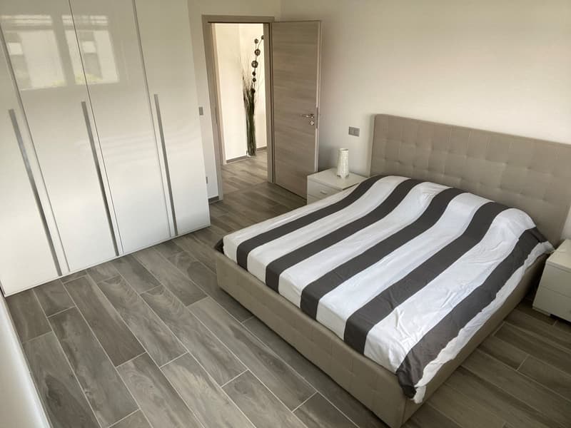 Bedroom with wooden floor, large bed, white walls, large wardrobe, nightstands, and an open door leading to another room.