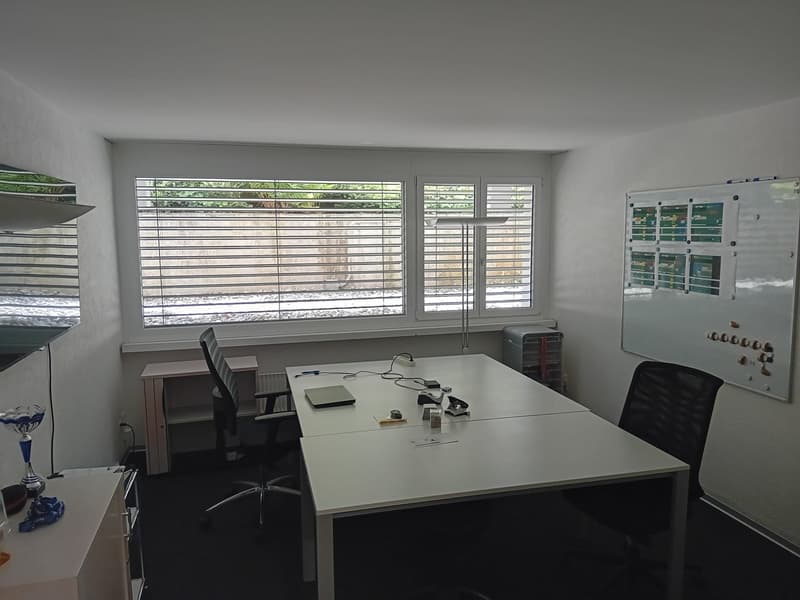 Office room with white walls, large window with blinds, white desk, two black office chairs, whiteboard, radiator, small table, lamp, trophy, and various office supplies.