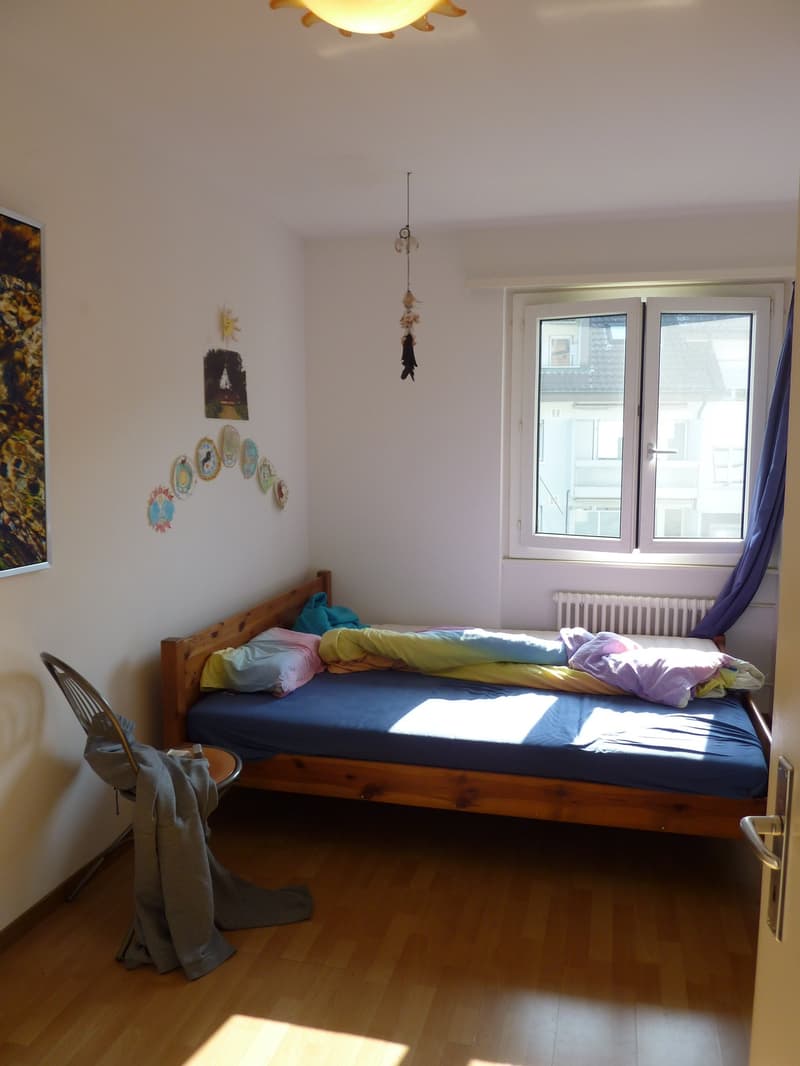 Bedroom with wooden bed, white walls, wooden floor, window with white curtains, chair, and a painting on the wall.