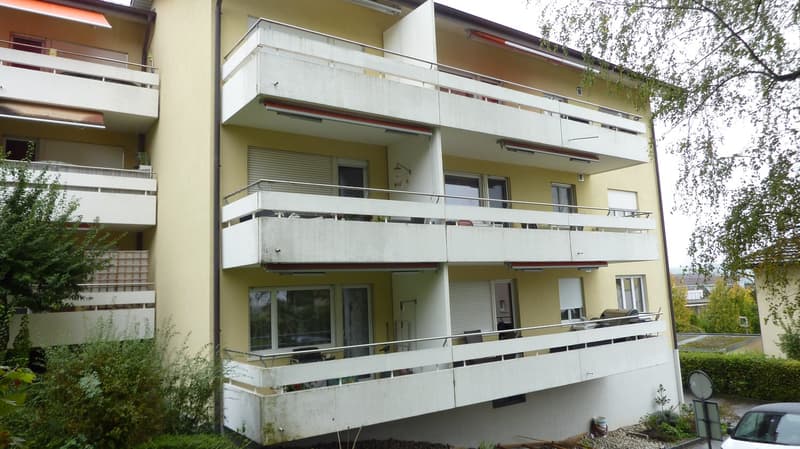 3 story building, yellow exterior, white balconies, multiple windows, metal railings, plants, trees