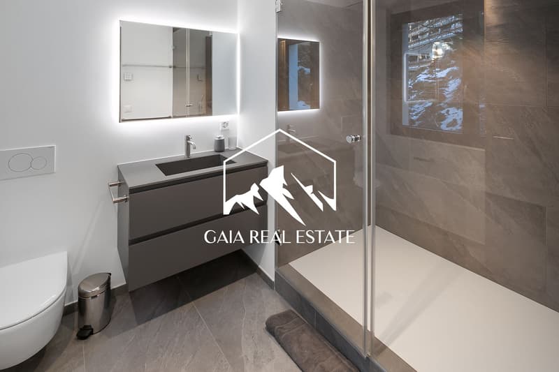 Modern bathroom with gray tile flooring, a large vanity with a black countertop, a mirror with built-in lighting, and a glass-enclosed shower area.
