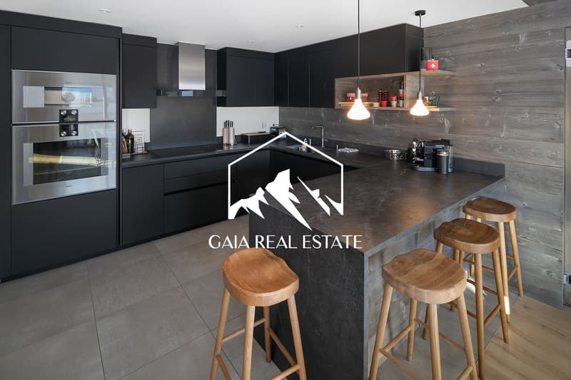 Modern, high-end kitchen with black cabinets, gray tile backsplash, wooden bar stools, and a Gaia Real Estate logo on the counter. The kitchen features a built-in oven, dishwasher, and other appliances.