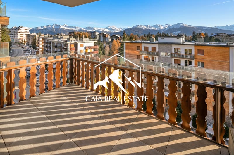 The image shows a balcony or terrace overlooking a scenic mountain landscape. The balcony has a wooden railing with intricate designs. The buildings in the background appear to be residential apartments or condos, with a mix of modern and traditional arch