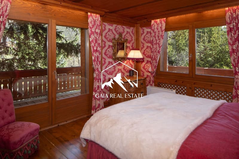 large bed, bedspread, two windows with balcony, red curtains, chair, wooden wall, wooden floor, lamp