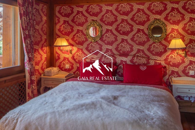 A bedroom with wallpaper, red color theme, a large bed, two nightstands, two wall lamps, two wall mirrors, a radiator, and a window.