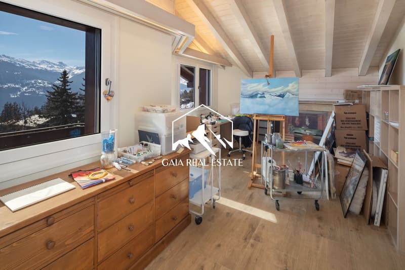 A room with wooden floor, wooden cabinet, desk, storage boxes, painting, easel, and a large window with a view of the mountains.