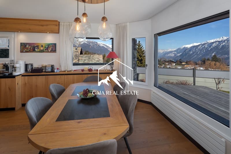 Dining room with wooden table, chairs, large windows, balcony, kitchen area with appliances, wooden cabinets, and a painting on the wall.