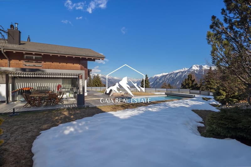 Modern chalet with wooden exterior, large windows, outdoor dining area, swimming pool, snowy landscape, mountain view